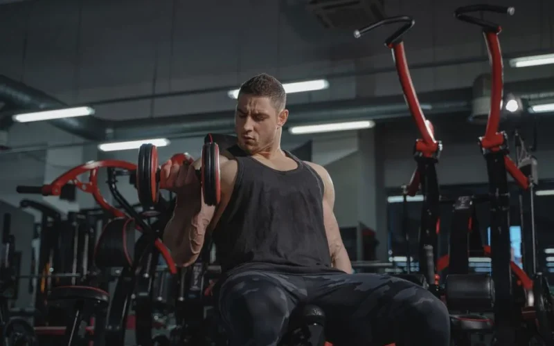 Athletic man performing dumbbell curls while sitting in a modern gym.