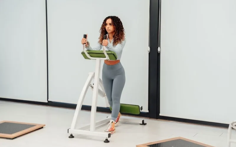 A woman with curly hair exercises on a machine in a bright gym.