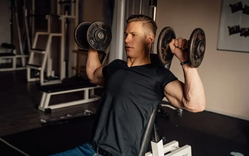 A focused man in a gym performing a dumbbell shoulder press, showcasing fitness and strength.