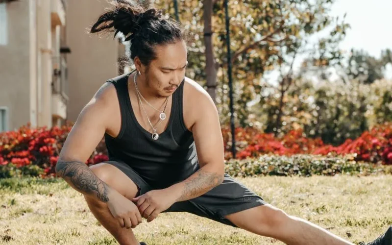 A tattooed man performing leg stretches outdoors in sunlight.