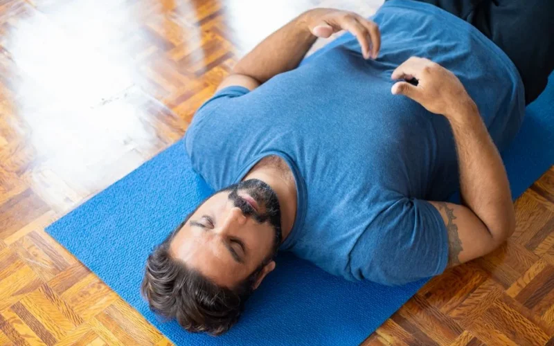 Adult man lying on a blue yoga mat indoors with eyes closed, focusing on relaxation and mindfulness.