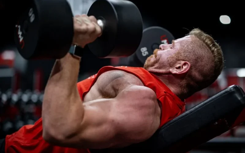 Side view of a muscular man lifting dumbbells on a bench in a gym, focusing on strength and effort.