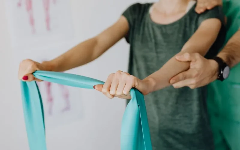 Crop unrecognizable orthopedist in uniform and wristwatch supporting shoulder and reached arm of faceless female patient with resistance band in doctor office