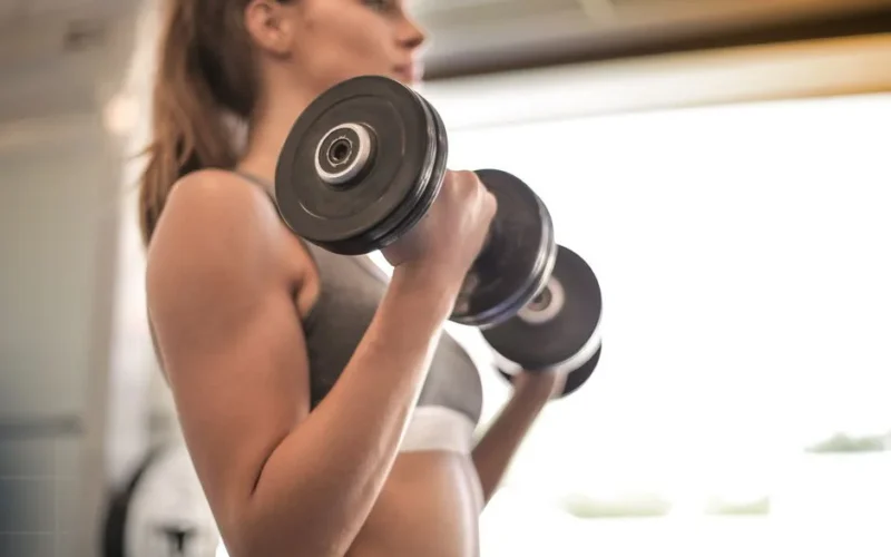 Focused woman lifting dumbbells in a bright gym, emphasizing strength and fitness.