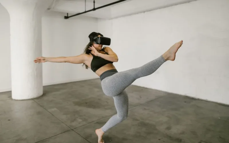 A woman practicing dance moves with a virtual reality headset in a minimalist indoor studio setting.
