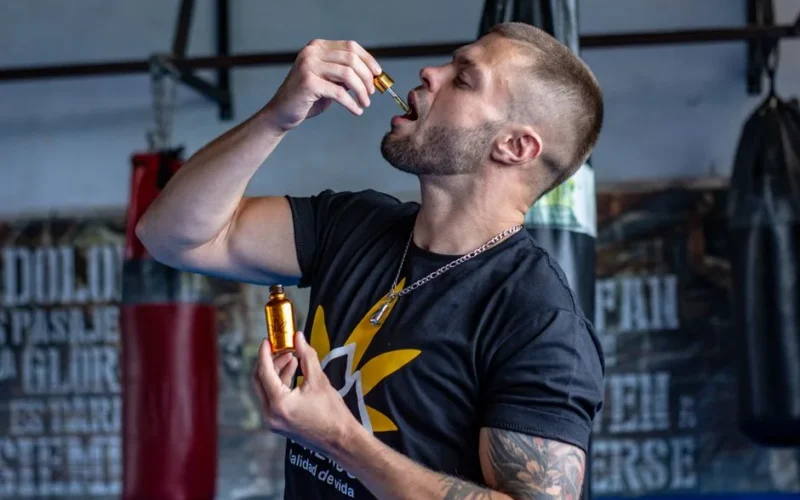 A muscular man in a gym takes a liquid supplement with a dropper.