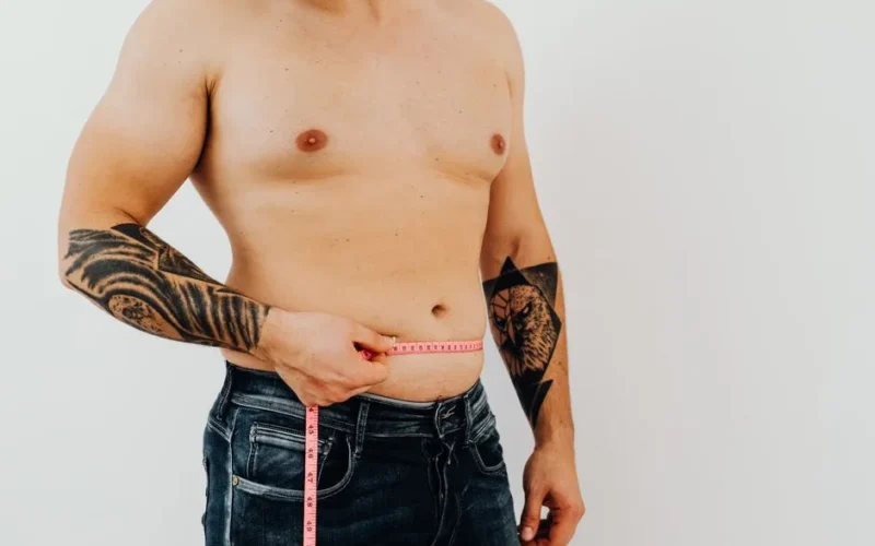Shirtless man using measuring tape to check waist size, showcasing tattoos for fitness concept.