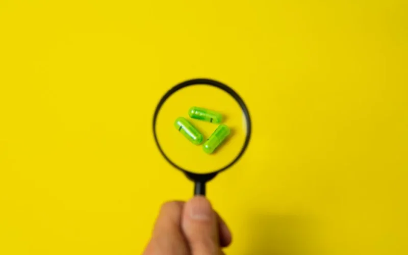Top view of crop unrecognizable person holding magnifier over green pills placed on bright yellow surface in studio