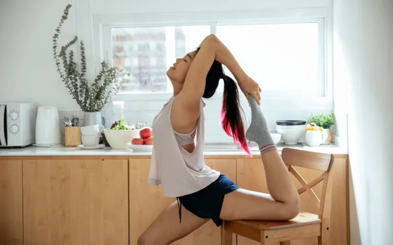 Side view of young slim Asian female stretching front surface of thigh using wooden chair while training Standing bow pose of Dandayamana Dhanurasana yoga asana at home