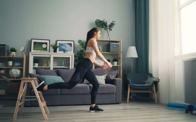 A woman in sportswear working out indoors using a chair for support, demonstrating leg exercises.