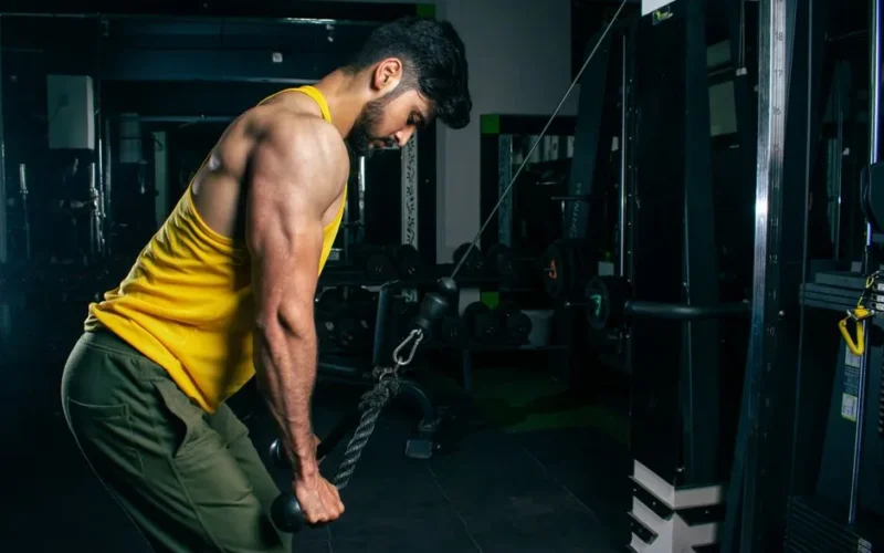 A fit man in a yellow tank top exercises using a cable machine in a gym setting.