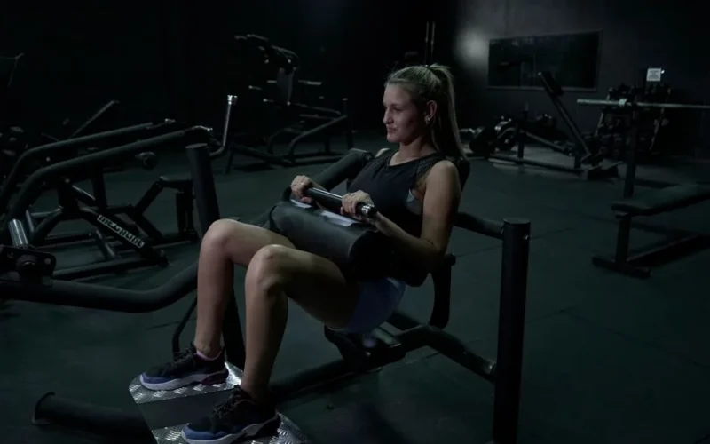 A woman performs leg exercises on a gym machine in a dimly lit environment.