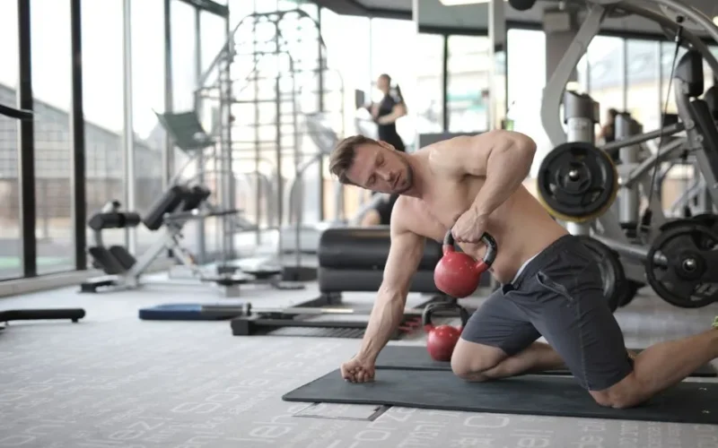 Shirtless man performing kettlebell exercise in a bright, modern gym space.