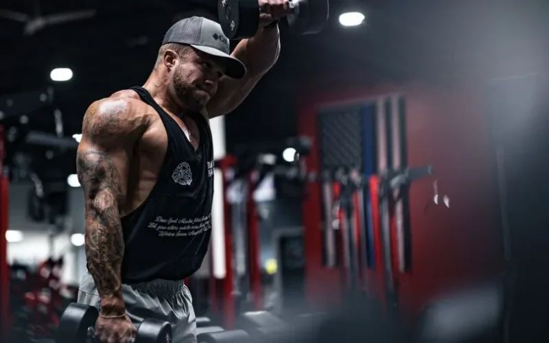 A muscular man with tattoos performs dumbbell exercises in a modern gym environment.