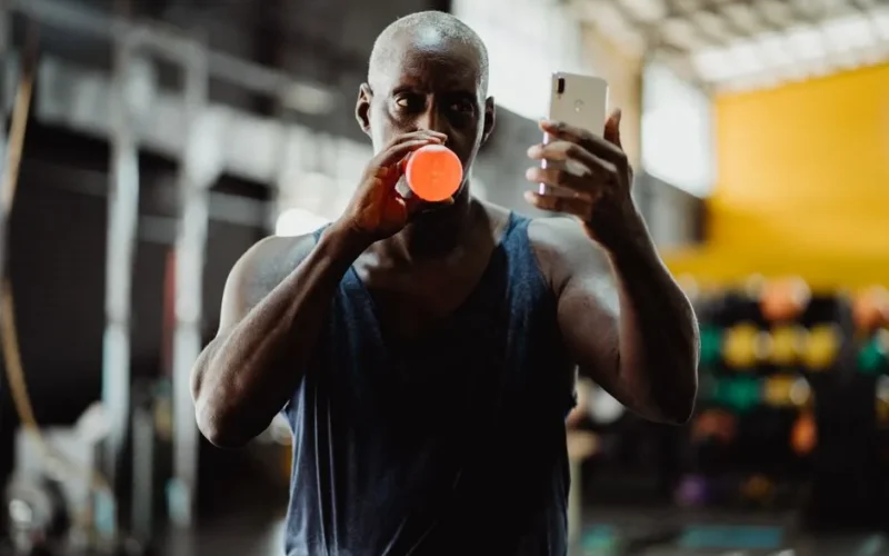 Athletic man drinking while checking smartphone in a modern gym environment.