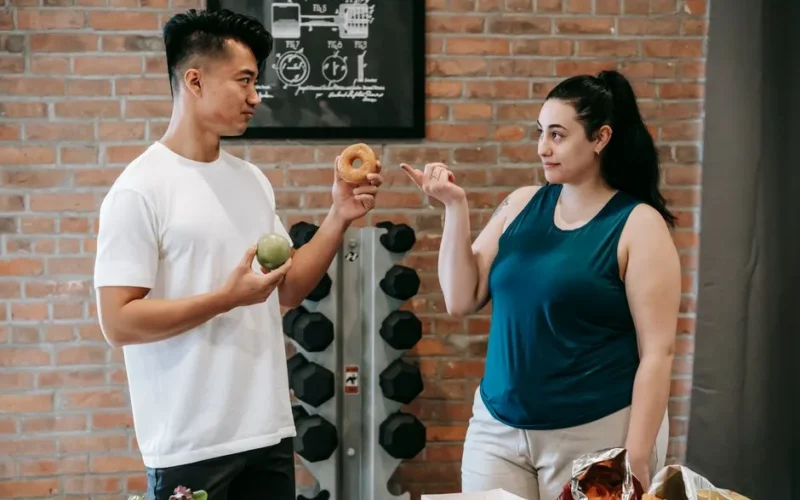 Side view of Asian personal instructor with apple and doughnut offering healthy food to plus size woman while standing in gym