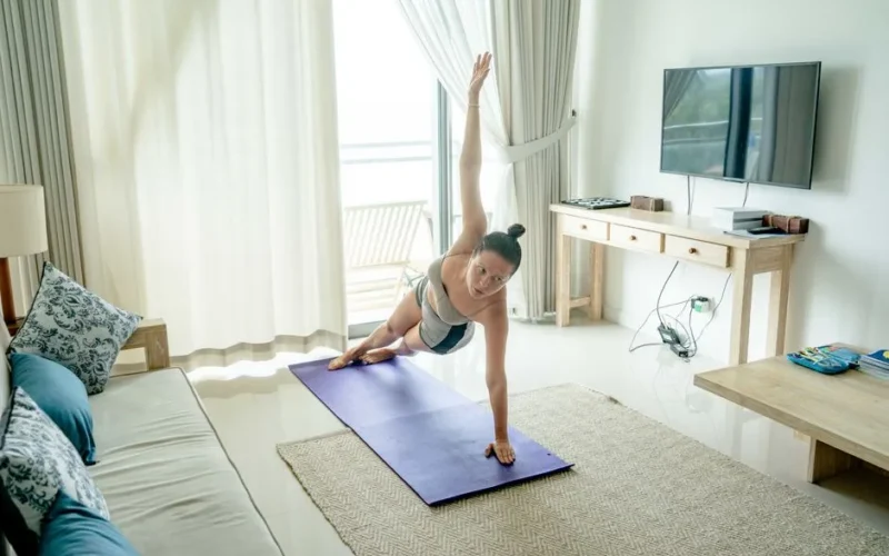 A woman practices yoga in a bright living room, performing a side plank on a mat.