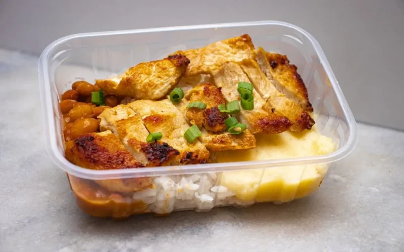 Grilled chicken with rice, beans, and greens in a clear takeout container.