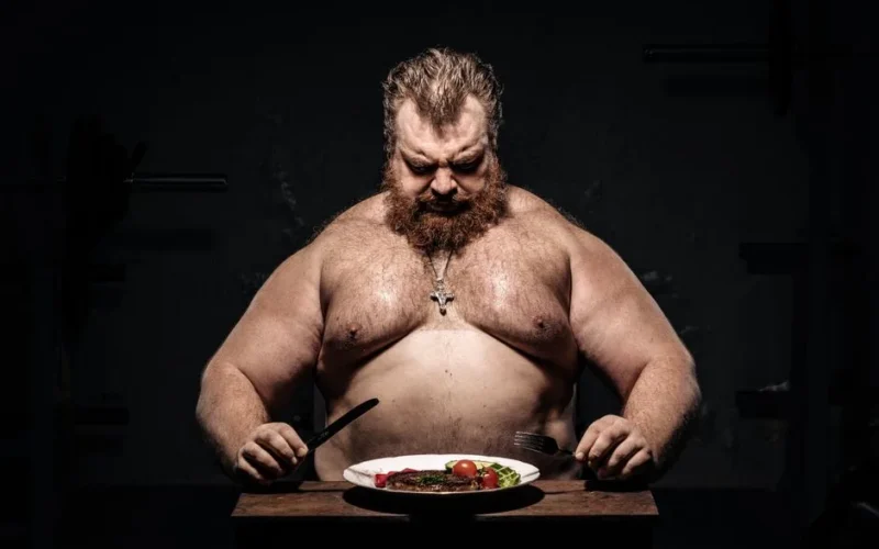 Bearded muscular man in dim light, preparing to eat steak with focus on fitness and nutrition.