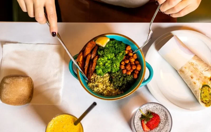 A vibrant vegan meal with fresh ingredients like broccoli, chickpeas, quinoa, served alongside a burrito and refreshing drinks.