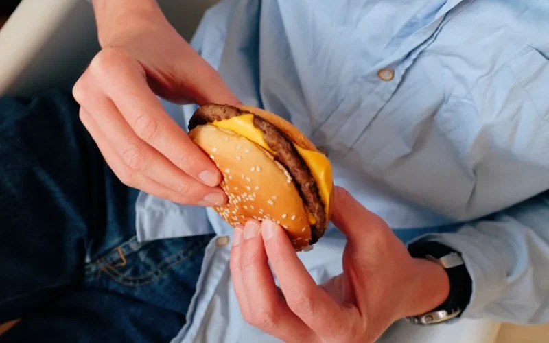 Unrecognizable male in blue shirt sitting on chair and holding delicious burger with cheese and cutlet in hands