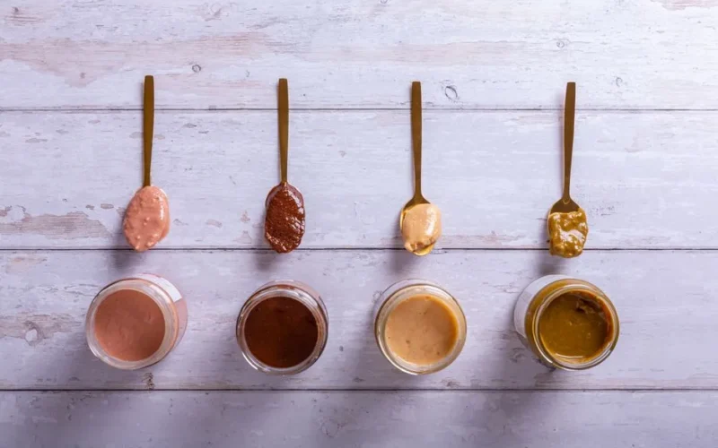 Top view of various nut butters in jars and spoons on a wooden surface.