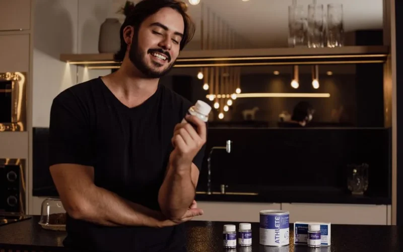 A smiling man in a kitchen holds a bottle of supplements, showcasing health and wellness.