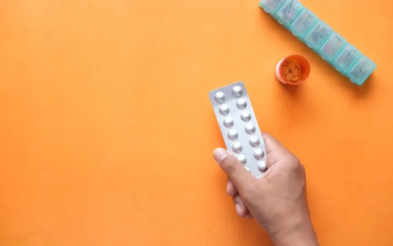 A hand holding a medicine blister near a pill organizer and orange pill bottle.