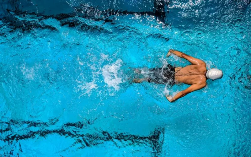 Top view of a swimmer wearing a cap, performing a front crawl stroke in a clear blue swimming pool.