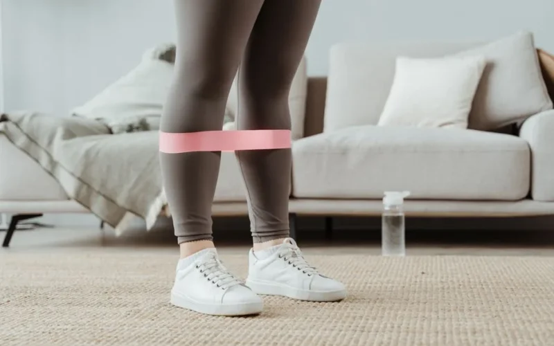 Close-up of a woman in sport leggings using a pink resistance band for leg exercises indoors.
