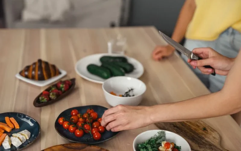 Hands slicing and arranging fresh vegetables and fruits on wooden table indoors.