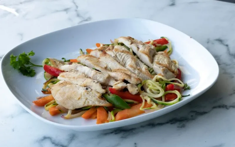 Healthy grilled chicken breast served on zucchini noodles and fresh vegetables.