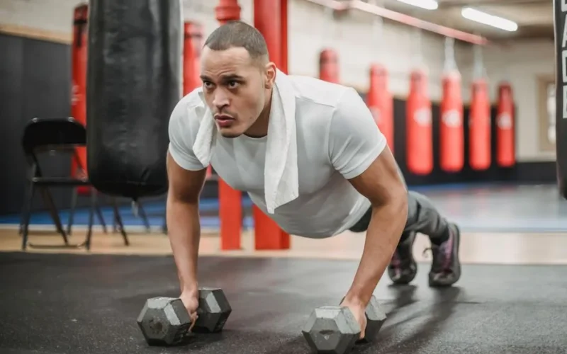 Full body powerful focused ethnic sportsman in activewear doing push up exercises with dumbbells during intense workout in contemporary gym