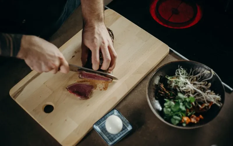A chef skillfully slices beef on a wooden board with fresh ingredients nearby.