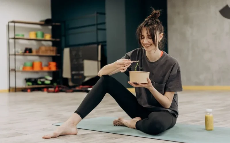 Young woman enjoying a healthy salad after exercising indoors. Promotes a balanced diet and fitness lifestyle.