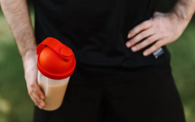 Close-up of a man holding a protein shake tumbler with a red lid outdoors, emphasizing fitness.