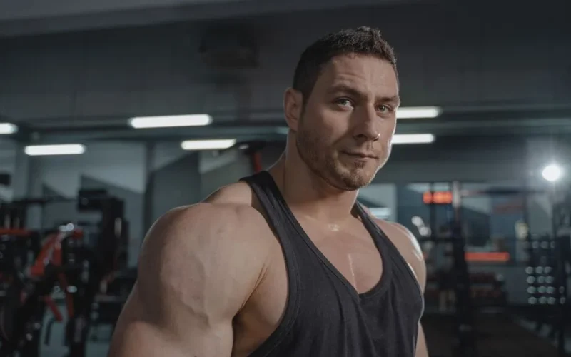 A muscular man in a black tank top posing confidently in a gym setting.