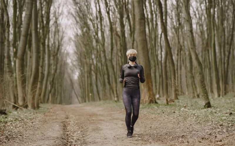 Full body woman in sportswear and disposable mask running along alley in park with headphones on neck during pandemic making workout