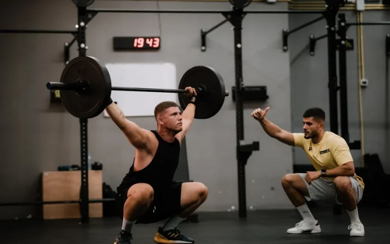 A muscular man performing a weightlifting squat with a coach guiding him indoors.