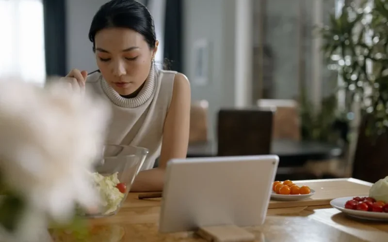Asian woman using tablet while preparing salad in modern kitchen.