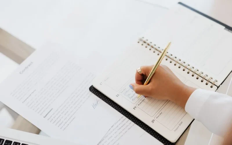 Close-up of a person writing in a planner next to a contract document on a desk.