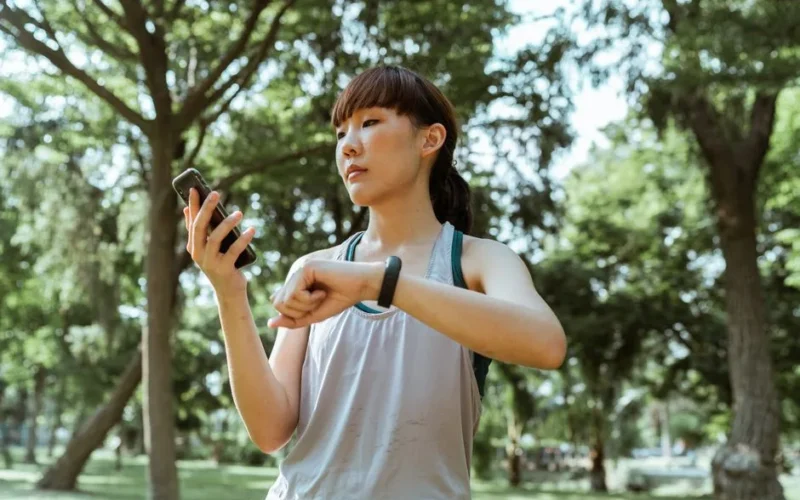 Positive concentrated young fit Asian sportswoman in activewear browsing smartphone and checking fitness bracelet while training in green sunny park