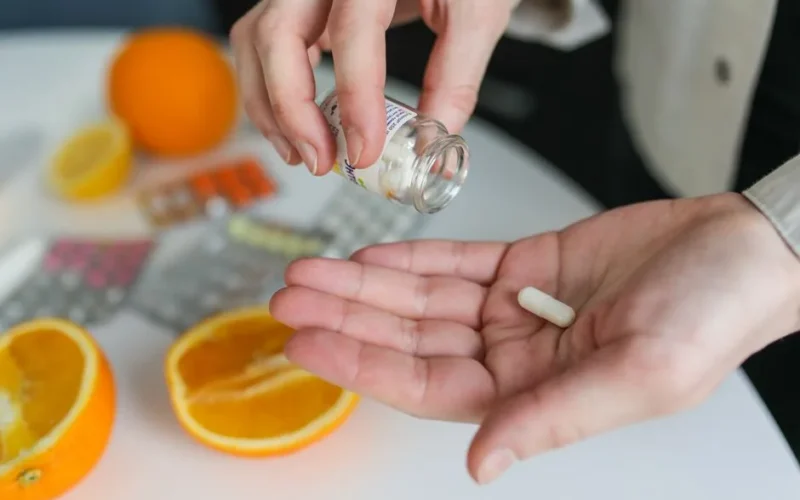 Close-up of a person taking a vitamin capsule with citrus fruit and medication on a table.