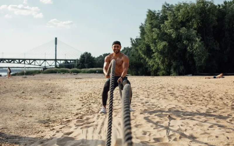 A muscular adult male with battle ropes exercises on a sunny beach by a bridge.