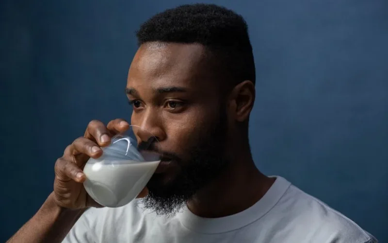 A serious man in a white t-shirt drinks milk from a glass against a dark blue background.