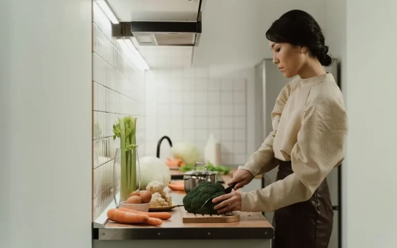 A woman chops broccoli on a kitchen counter with various fresh vegetables around.
