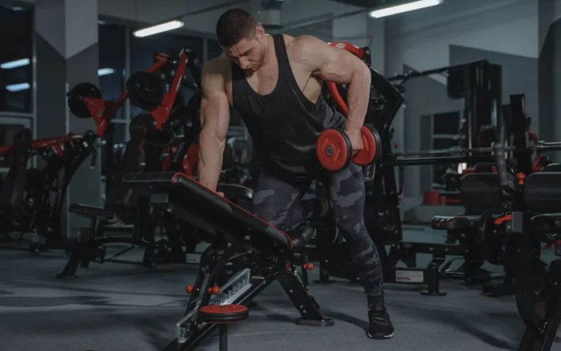 A strong, muscular man lifting dumbbells in a modern gym environment.
