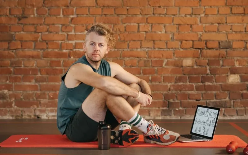 A man sits on an exercise mat with a laptop showing workout routines, suggesting a fitness regimen.