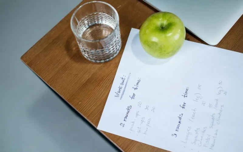 A green apple, glass of water, and workout plan on a wooden desk symbolize healthy living.
