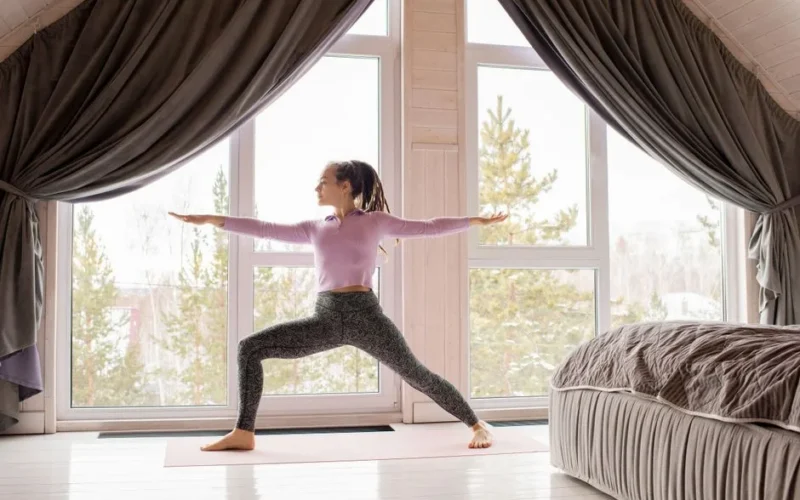 Adult woman doing yoga in a peaceful bedroom with large windows, focusing on stretching and mindfulness.